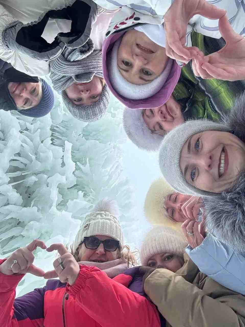 Группа людей в зимней одежде делает сердечки руками на фоне снега.