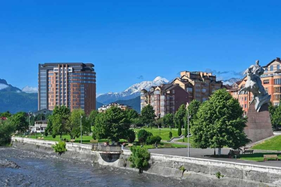 Панорама города с рекой и горами Городская панорама с рекой, зеленью и скульптурой на фоне гор.